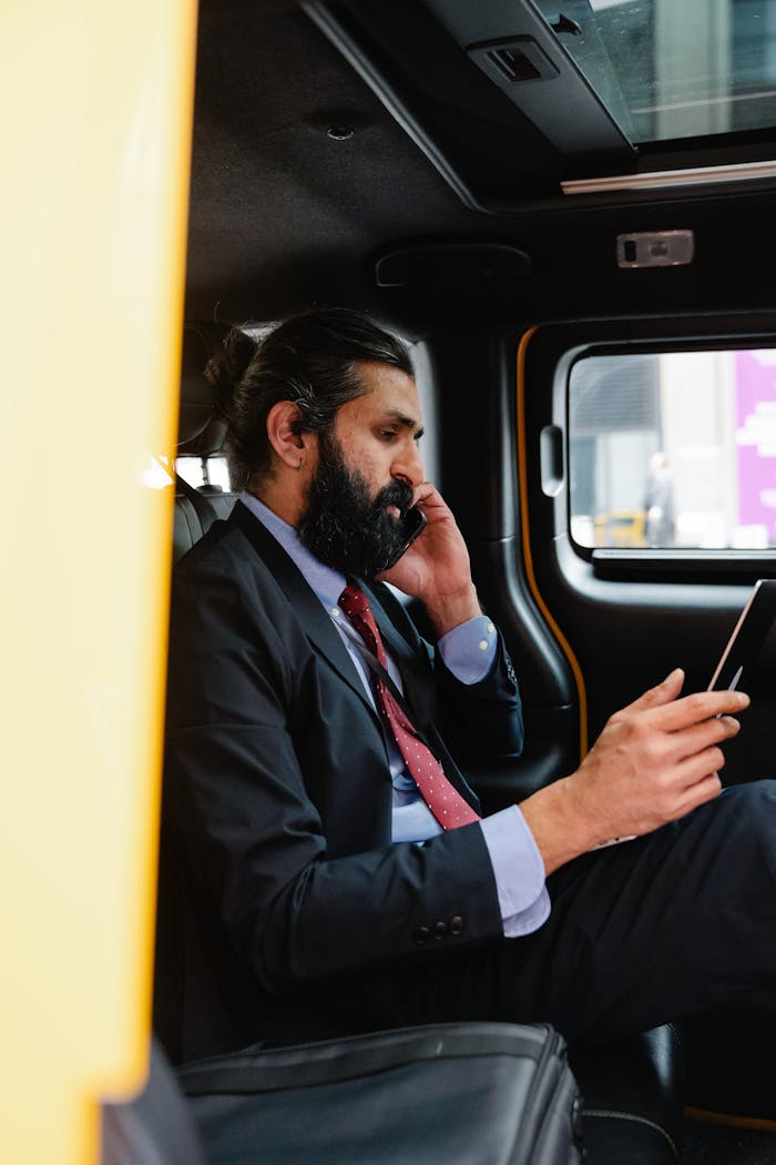 Bearded businessman multitasking on phone and tablet in car interior.